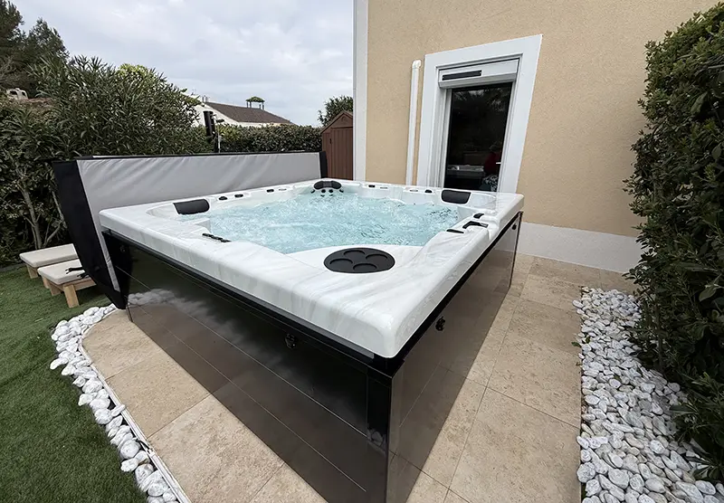 Zoom sur les finitions d'un spa 5 places Napoli Luxe installé par Azur Spa Design. Intégration parfaite entre une terrasse moderne, des bordures en galets blancs et une façade de maison, offrant un espace de balnéothérapie à domicile dans le Var.