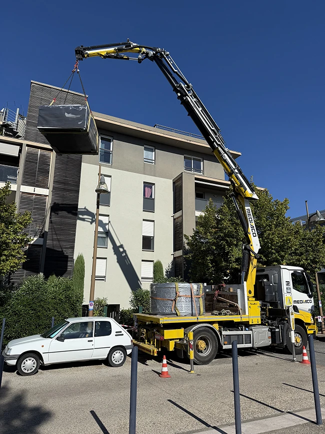 Un camion-grue jaune soulève un spa emballé au-dessus d'une voiture blanche stationnée, en vue d'une installation sur un toit-terrasse d'immeuble à Fréjus, sous un ciel bleu