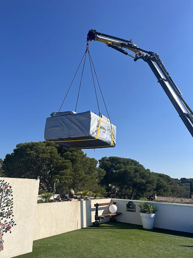 Un spa sous housse de protection est suspendu dans le vide au bout du bras d'une grue au-dessus d'un toit-terrasse avec gazon synthétique, banc, et pots de fleurs à Saint-Raphaël.